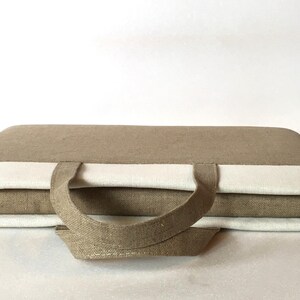 May include: A brown linen laptop bag with white trim and a top handle. The bag is closed and the handle is folded down.