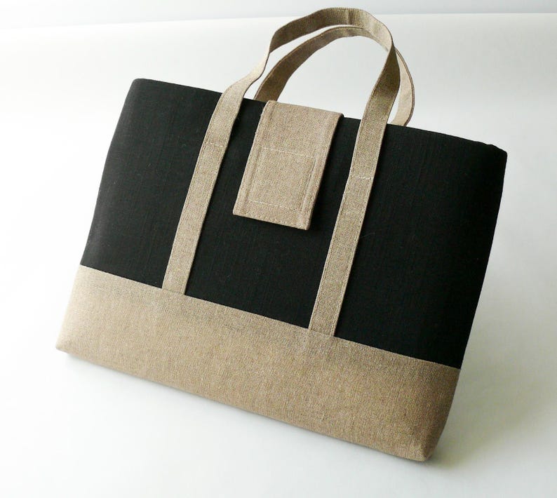 May include: A rectangular tote bag with a black upper section and a tan lower section. The bag has tan handles and a small tan pocket on the front. The bag is made of a textured fabric and is sitting on a white surface.