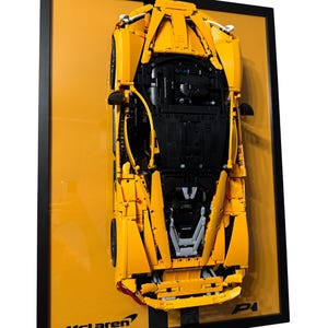 May include: A framed yellow McLaren model car, viewed from above, against a yellow background. The car is constructed from interlocking blocks, with black detailing. The frame is black, and the McLaren logo is visible at the bottom left.