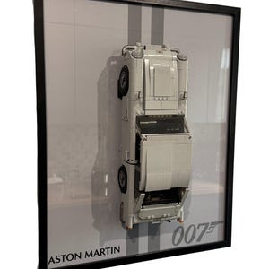 Può includere: Opera d'arte incorniciata con un'auto modello Aston Martin bianca costruita con mattoncini giocattolo. L'auto è posizionata verticalmente su uno sfondo grigio con due strisce grigie verticali. La cornice è nera, con i testi "ASTON MARTIN" e "007".