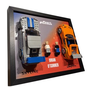May include: A framed display featuring two toy cars and two minifigures. A silver and blue Skyline and an orange Supra are mounted alongside figures labeled Brian O'Conner. The background includes the Fast & Furious logo and text.