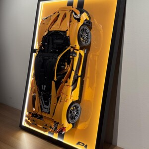 May include: A framed display featuring a yellow and black model sports car. The car is set against an orange background with a black stripe. The frame has a black border with a built-in light.