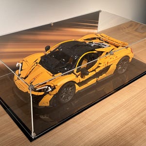 May include: A yellow and black model sports car constructed from interlocking plastic bricks, displayed inside a rectangular clear acrylic protective case with a black base, featuring detailed body panels, dark wheels, and a rear spoiler.