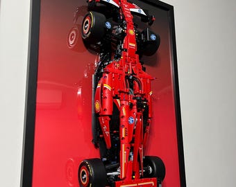 Könnte beinhalten: Ein rotes Lego Ferrari Formel-1-Rennwagen wird in einem schwarzen Rahmen vor rotem Hintergrund ausgestellt. Das Auto schwebt in der Luft, wobei die Vorderräder zum Betrachter zeigen. Das Auto trägt das Ferrari-Logo und die Nummer 5 an der Seite.