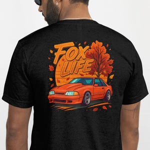 May include: Black t-shirt featuring a graphic of an orange car with the text "Fox Life" and an autumn-themed background. The design includes a stylized car, a tree, and a sun-like circle. The shirt is made of a soft material.