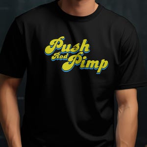 May include: Black t-shirt with the text "Push Rod Pimp" in yellow script with a blue outline. The shirt is short-sleeved and appears to be made of a soft material. The person wearing the shirt has a red watch on their wrist.