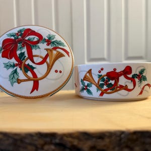 May include: A white ceramic bowl and lid set with a festive Christmas design. The design features a red bow, gold horn, holly, and red berries. The rim is trimmed with a gold and green border.
