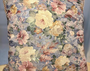 Puede incluir: Cojín cuadrado floral con fondo azul y flores amarillas, rosas y moradas. Cuenta con un ribete de cuerda alrededor de los bordes.