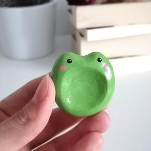 Peut inclure: Un petit objet vert en forme de grenouille, tenu dans une main. La grenouille a des yeux noirs et des joues roses. L'objet semble être en céramique peinte ou dans un matériau similaire. L'arrière-plan comprend une surface blanche et une plante en pot.