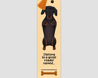 Peut inclure: Marque-page rectangulaire avec une illustration de teckel. Le chien est noir et marron, debout. Le marque-page porte le texte "I belong to a great reader named..." et un os. Une gland orange est suspendu en haut.