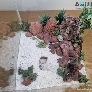 Top-down view of an aquarium setup featuring the Riptide Aquarium Cave STL file surrounded by sand, rocks, moss, and plants, showcasing a natural aquascaping design. Ideal for fish or reptiles.