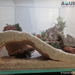 Side view of the Riptide Aquarium Cave STL file in a natural aquarium setting with sand, rocks, moss, and plants, showcasing its spacious design for fish or reptiles. Suitable for 3D printing.