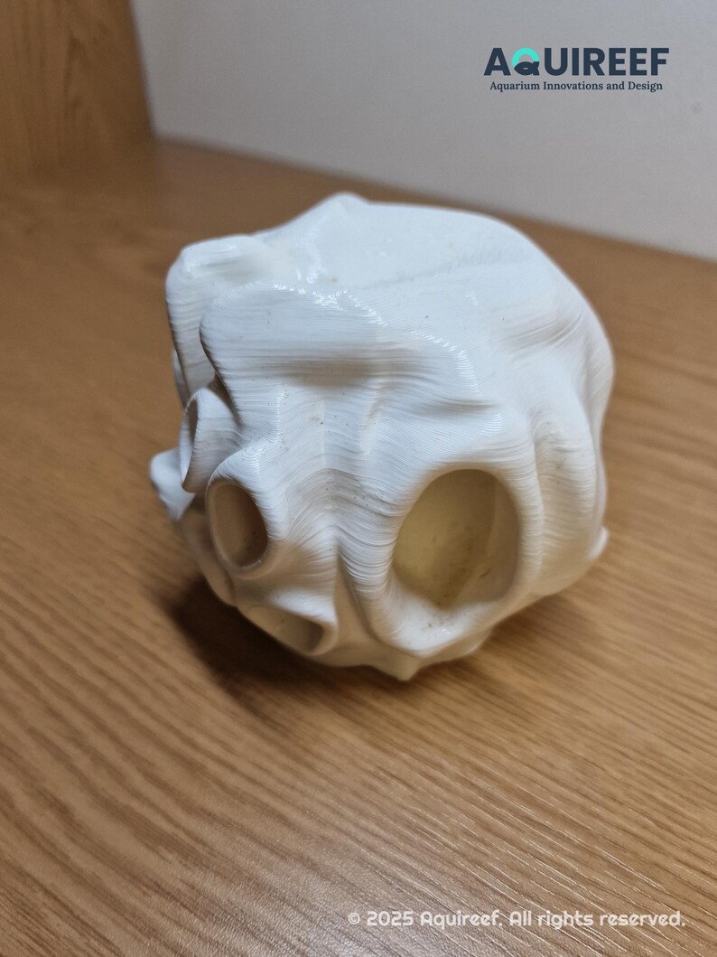 A side view of a white 3D-printed aquarium cave with smooth curves and multiple oval openings. Placed on a wooden surface, the design showcases an organic, flowing structure ideal for fish to explore and hide.