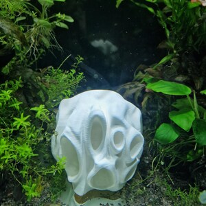A white 3D-printed aquarium cave with multiple oval-shaped openings sits on a gravel substrate surrounded by green aquatic plants. The design provides hiding spots and exploration spaces for fish in an underwater environment.