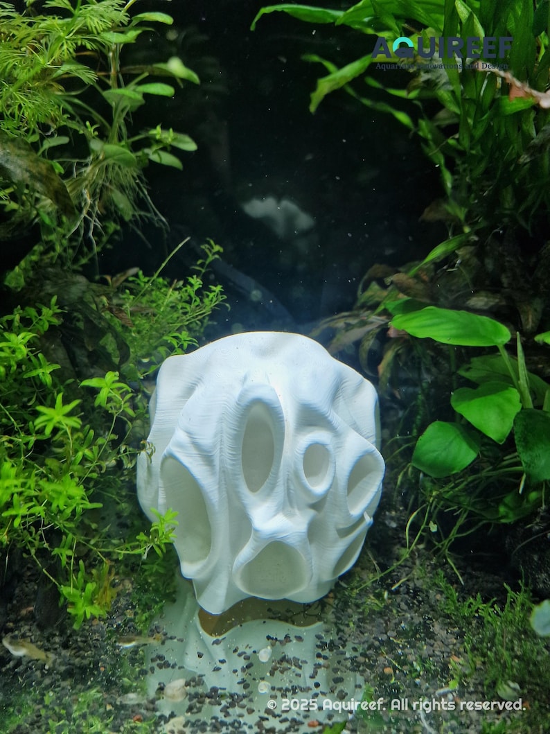 A white 3D-printed aquarium cave with multiple oval-shaped openings sits on a gravel substrate surrounded by green aquatic plants. The design provides hiding spots and exploration spaces for fish in an underwater environment.