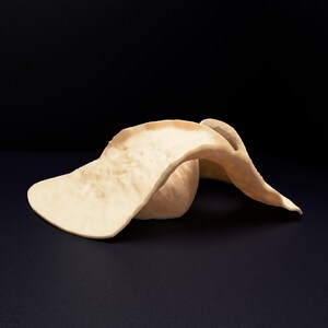 Op de afbeelding: Een sculpturaal object gemaakt van gebroken wit materiaal met een gladde, golvende en gevouwen vorm. Het stuk heeft organische, gebogen randen en een matte textuur, wat een vloeiende vorm cre&euml;ert die op een plat oppervlak rust.