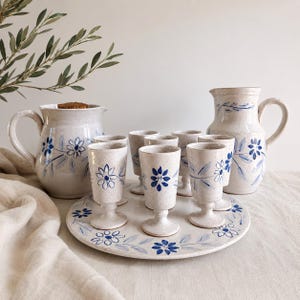 May include: A ceramic drinkware set featuring a pitcher, a serving tray, and stemmed glasses. The set is off-white with blue floral designs. The pitcher and glasses have handles. The tray is round and decorated with blue flowers.