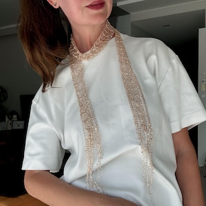 May include: A long, thin, rose gold metallic scarf with an open, delicate, net-like crochet texture. The scarf is draped around the neck, with two long, slender ends hanging down over a plain, short-sleeved white t-shirt, creating a layered look.