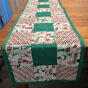 May include: A Christmas-themed table runner with a patchwork design. The runner features alternating blocks of green fabric and fabric with red and white candy cane stripes and gnome illustrations. The edges are trimmed with green fabric.