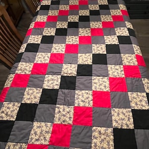 May include: A handmade quilt with a patchwork design. The quilt features a pattern of alternating squares in red, black, gray, and a beige fabric with a repeating pattern of small figures. The quilt is spread out on a table.