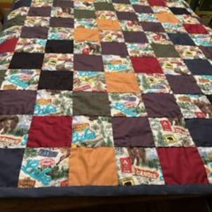 May include: A patchwork quilt with a variety of square patterns in shades of red, orange, blue, green, and brown. Some squares feature outdoor-themed prints, while others are solid colors. The quilt is displayed on a bed.