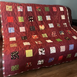 May include: A burgundy patchwork quilt with a plaid border. The quilt features numerous square patches in various patterns, including floral, striped, and holiday-themed designs. The quilt is draped over a brown leather couch.