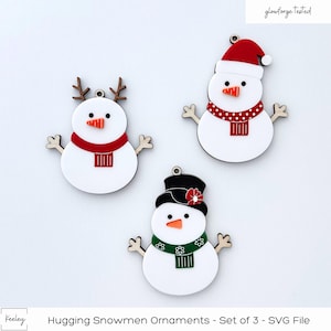 May include: Set of three wooden snowman ornaments. Each snowman has a different hat and scarf. The snowman on the left has antlers, the snowman in the middle has a red Santa hat, and the snowman on the right has a black top hat with a red flower.