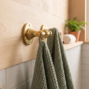 May include: A polished brass double hook mounted on a light wood panel, featuring a rounded base and two protruding pegs. Two sage green waffle-textured towels are draped over the hooks, hanging down against a white tiled wall surface.