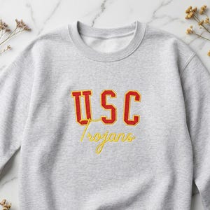 Könnte beinhalten: Ein hellgraues Sweatshirt mit Rundhalsausschnitt, das den Schriftzug USC in fetten roten Blockbuchstaben mit gelber Umrandung zeigt, platziert über dem Wort Trojans, das in gelber Schreibschrift auf der Brust gestickt ist.