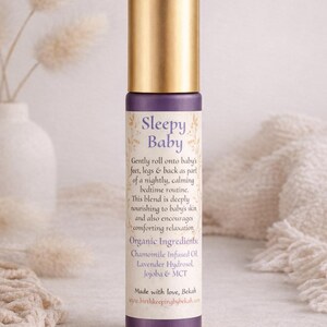 May include: A purple and gold essential oil roller bottle labeled "Sleepy Baby." The label lists organic ingredients and instructions for use. The bottle is set against a soft, neutral background with decorative elements.