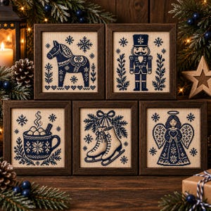 May include: Five framed cross-stitch artworks featuring dark blue thread on a light beige fabric. The designs include a Dala horse, a nutcracker, a mug of hot cocoa, ice skates, and an angel, all surrounded by snowflake and botanical motifs.