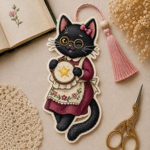 Può includere: Un segnalibro a punto croce a forma di gatto nero con occhiali tondi dorati e un fiocco rosa. Il gatto indossa un vestito bordeaux con un grembiule floreale e tiene in mano un piccolo telaio da ricamo con una stella gialla. Una nappa rosa è attaccata.