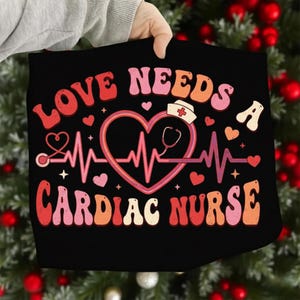 Peut inclure: T-shirt noir avec l'inscription "LOVE NEEDS A CARDIAC NURSE" en police rétro. Le motif comprend un cœur, un stéthoscope et un chapeau d'infirmière. Le design est dans les tons de rouge, rose et orange.