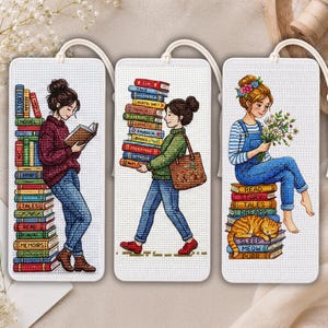 Può includere: Tre segnalibri rettangolari in tessuto bianco con illustrazioni a punto croce. Il primo mostra una persona che legge accanto a una pila di libri. Il secondo mostra una persona che trasporta libri e una borsa. Il terzo mostra una persona con fiori seduta su libri con un gatto.