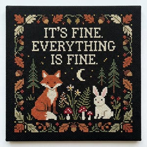 May include: A square cross-stitch embroidery on black fabric featuring the text IT'S FINE. EVERYTHING IS FINE. in white. The design includes a red fox, a white rabbit, pine trees, mushrooms, a crescent moon, and a border of autumn leaves and acorns.