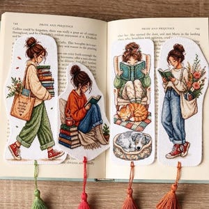 Puede incluir: Cuatro marcadores bordados con borlas. Cada marcador presenta una ilustración diferente de una persona leyendo, con libros, un gato y un perro. Los marcadores están colocados en las páginas de un libro.