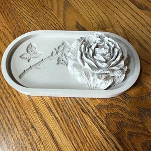 May include: An oval-shaped light gray tray featuring a three-dimensional rose flower sculpture on the right side and a recessed, etched design of a rose stem with a single leaf on the left side. The tray has a smooth, matte finish throughout.
