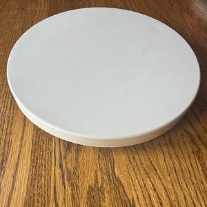 Pode incluir: Uma bandeja de servir circular, plana e branca, com uma superfície lisa e mate e uma borda vertical baixa. A bandeja apresenta um design limpo e uniforme, proporcionando uma plataforma estável e nivelada para vários itens.