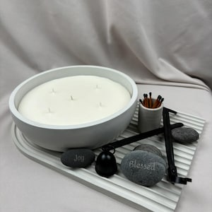 May include: White concrete candle bowl with four wicks, surrounded by black sticks, a small black metal bell, and three gray stones with the words "Joy" and "Blessed" inscribed on them.