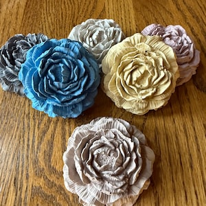 May include: A collection of six flower-shaped decorative objects made of a textured material. The set includes individual pieces in shades of bright blue, pale yellow, light gray, dark gray, and soft lavender, each featuring layered petals.