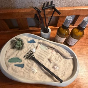 May include: A miniature zen garden with sand, decorative rocks, shells, and a small rake. Two brown glass spray bottles are labeled "Cabana" and "Seaside Retreat". A small concrete pot holds zen garden tools.