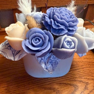 May include: A floral arrangement featuring soap flowers in shades of blue and cream. The arrangement includes roses and other flower shapes, accented with dried grasses and feathers, presented in a light blue container with a patterned ribbon.
