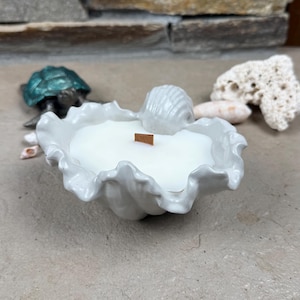 May include: A white candle in a glossy, ruffled ceramic shell-shaped container. The candle features a flat, rectangular wooden wick centered in the white wax. The shell vessel has a scalloped edge and a small, molded shell detail on the rim.