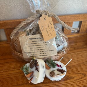 May include: A gift set of fire starters in a woven basket, wrapped in clear cellophane and tied with twine. The set includes heart-shaped and round wax fire starters decorated with pinecones and herbs.