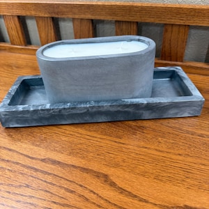 May include: A gray concrete candle in an oval shape, resting on a matching rectangular tray. The candle has a white wax top. The set is displayed on a wooden surface.
