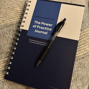 Puede incluir: Una libreta de espiral azul y blanca con el título "The Power of Practice Journal" y el texto "Scientifically Designed Journal to Facilitate Skill Acquisition". Un bolígrafo negro está sobre la libreta.