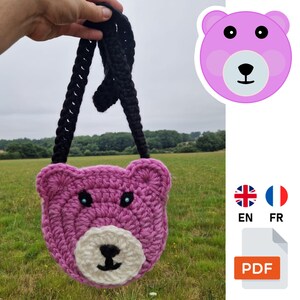 May include: A handmade, crochet bear-shaped bag in shades of pink, black, and white. The bag features a bear face design with blue eyes and a black nose. The bag has a black strap and is held up against a green field background.