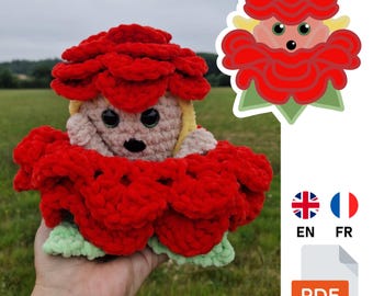Op de afbeelding: Handgemaakte gehaakte pop in de vorm van een rode bloem met een lichtbruin gezicht, zwarte ogen en geel haar. De pop wordt in een hand gehouden. Een cartoon bloem illustratie en PDF icoon zijn ook zichtbaar.