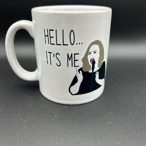 May include: White ceramic mug with the text "HELLO... IT'S ME" and an illustration of a singer. The mug has a curved handle and a black and white graphic of a singer holding a microphone.
