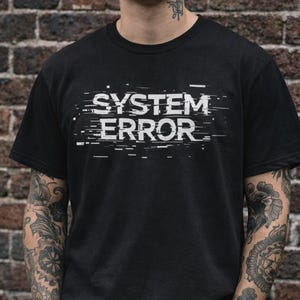 Op de afbeelding: Zwart T-shirt met de witte tekst "SYSTEM ERROR" in een verstoorde lettertype. Het shirt met korte mouwen heeft een ronde hals. De persoon staat voor een bakstenen muur.